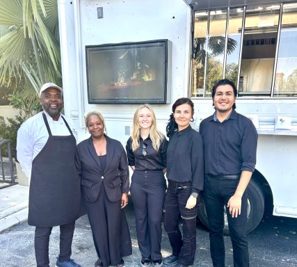 Five diverse individuals stand together outside a food truck, smiling and dressed in black attire.