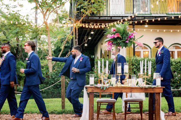 Groom and groomsmen in blue suits walking past a decorated outdoor reception setup.