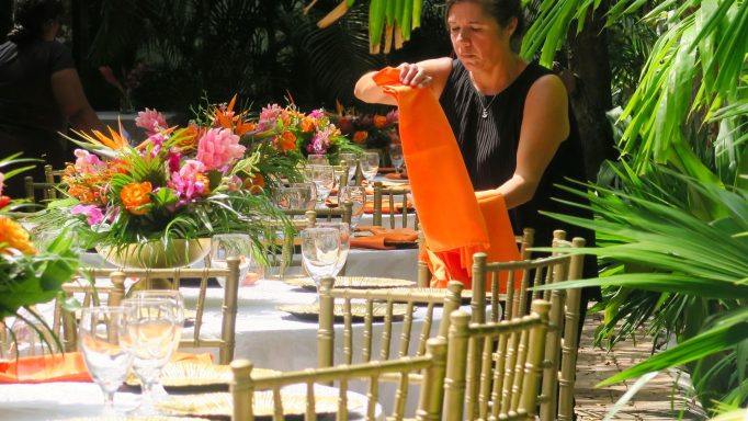 A woman arranges an orange napkin on a beautifully set dining table surrounded by greenery.