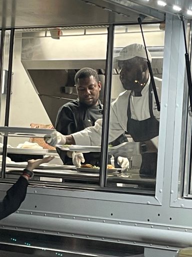 Two chefs serve food from a window in a food truck.