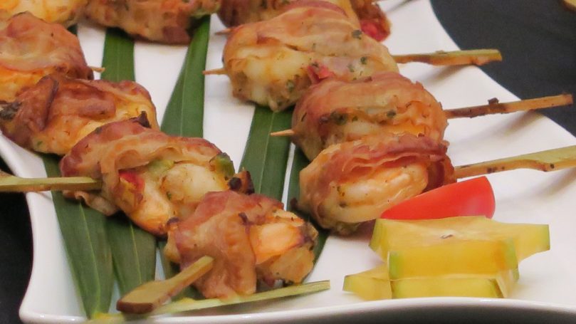 Grilled skewers of shrimp wrapped in bacon on a platter with garnish.